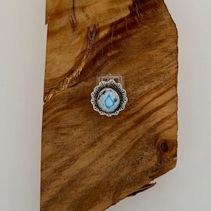 Könnte beinhalten: Ein Silberring mit einem zentralen, ovalen, hellblauen Stein mit dunklen Einschlüssen, umgeben von einem gewellten Metallrand mit eingravierten Liniendetails. Der Ring ist auf einem durchsichtigen Kunststoffständer befestigt.
