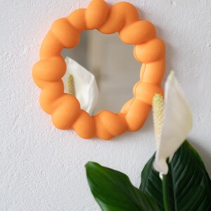 May include: An orange, round mirror with a unique, textured frame. The frame is made of a series of connected, rounded shapes. The mirror is hanging on a white wall, with a plant with green leaves and white flowers in the foreground.