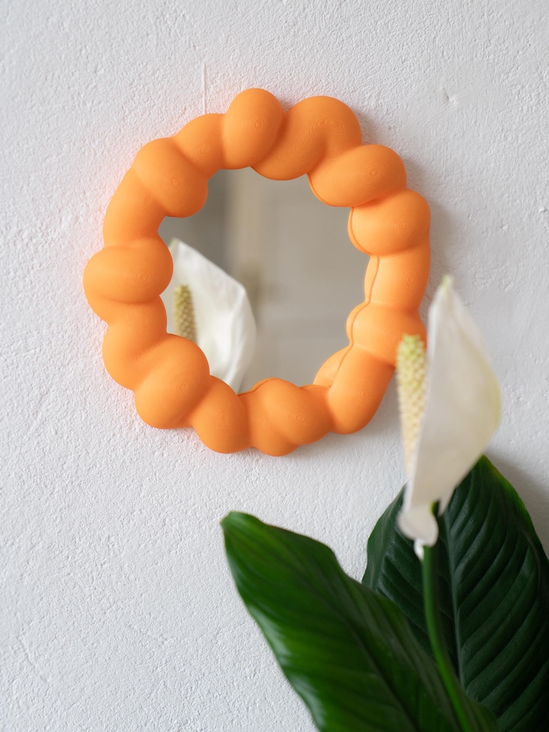 May include: An orange, round mirror with a unique, textured frame. The frame is made of a series of connected, rounded shapes. The mirror is hanging on a white wall, with a plant with green leaves and white flowers in the foreground.