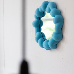 May include: A round, light blue mirror with a unique, cloud-like frame. The frame is made of rounded, textured shapes. The mirror is mounted on a white wall, reflecting a glimpse of the outdoors. The overall aesthetic is modern and playful.