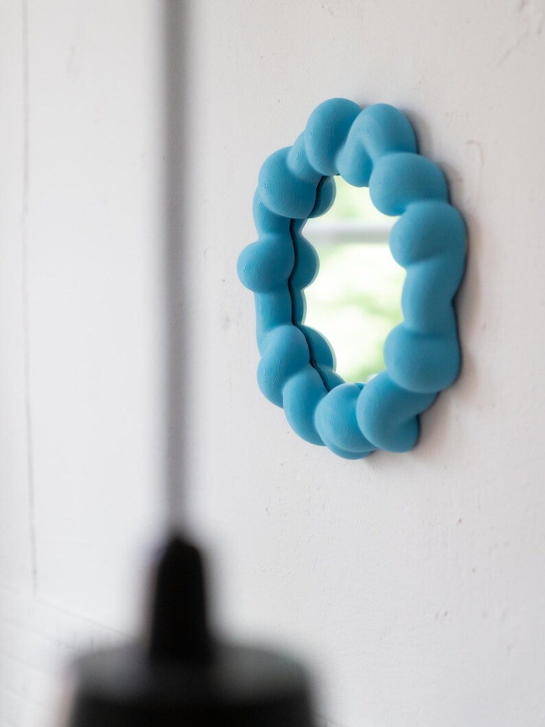May include: A round, light blue mirror with a unique, cloud-like frame. The frame is made of rounded, textured shapes. The mirror is mounted on a white wall, reflecting a glimpse of the outdoors. The overall aesthetic is modern and playful.