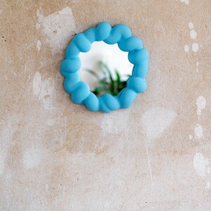 May include: A small, round mirror with a light blue, cloud-like frame. The mirror is mounted on a textured, beige wall. The reflection shows a glimpse of green foliage.