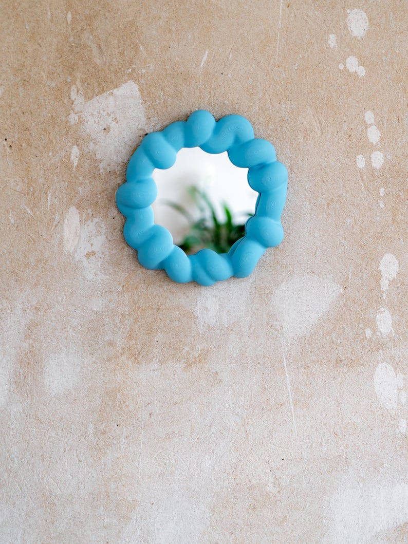 May include: A small, round mirror with a light blue, cloud-like frame. The mirror is mounted on a textured, beige wall. The reflection shows a glimpse of green foliage.
