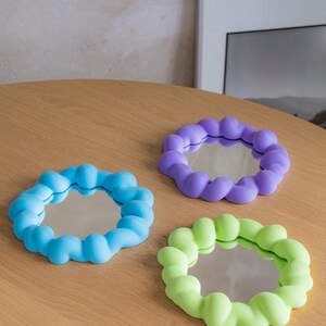 May include: Three small, round mirrors with colorful, bubble-like frames. One is light blue, another is purple, and the third is lime green. The mirrors are displayed on a light brown wooden surface.