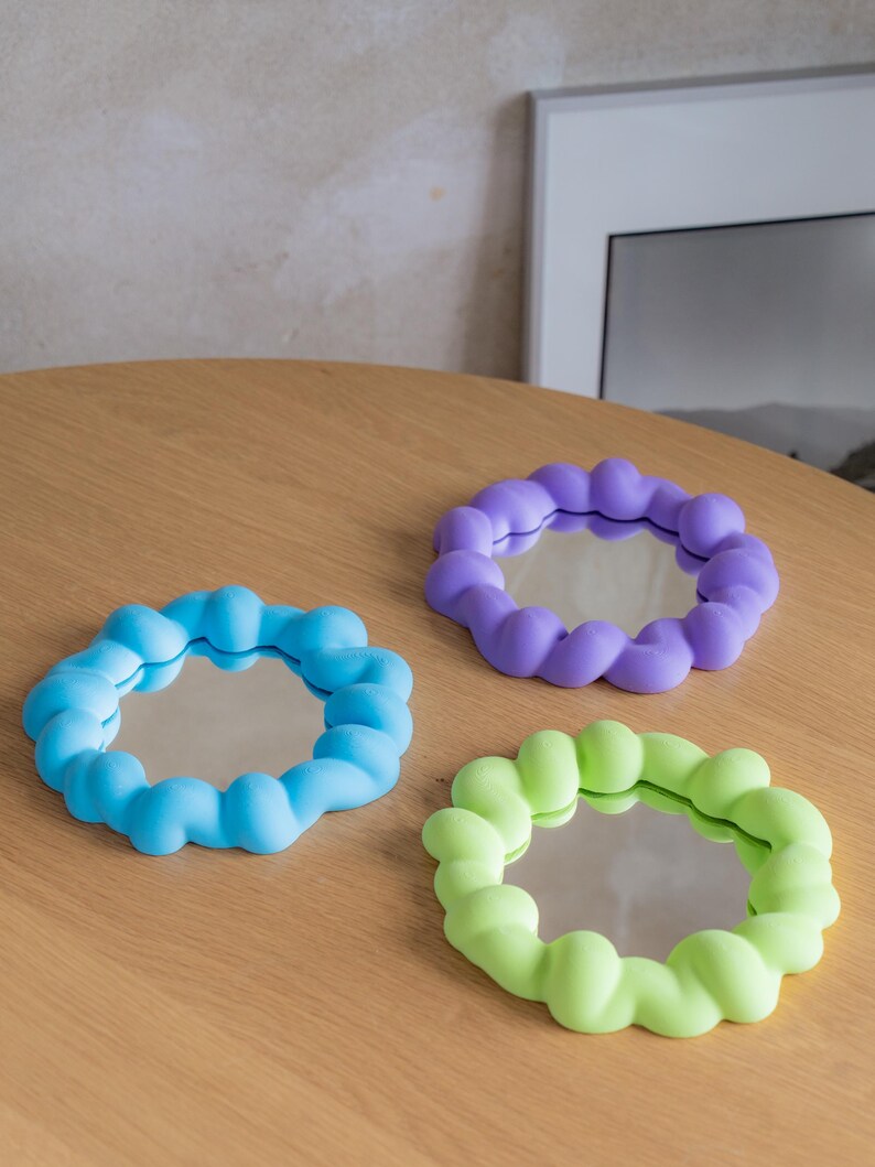 May include: Three small, round mirrors with colorful, bubble-like frames. One is light blue, another is purple, and the third is lime green. The mirrors are displayed on a light brown wooden surface.