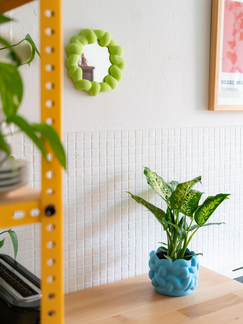 May include: A small, round mirror with a lime green, bubble-like frame is mounted on a white wall. A blue, textured planter holds a green and white plant on a wooden surface. A yellow metal shelf is visible in the background.