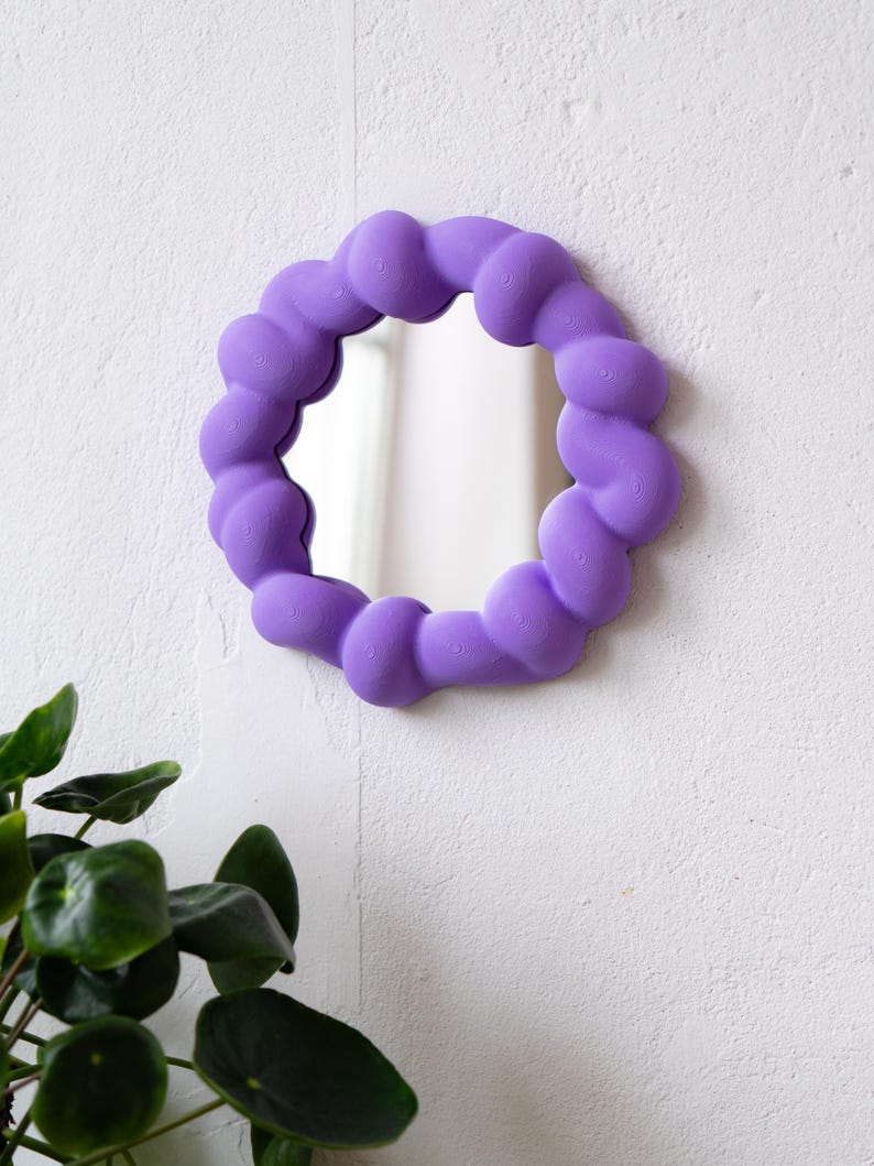 May include: A round, purple mirror with a unique, cloud-like frame. The mirror is mounted on a white wall, with a small green plant visible in the lower left corner. The frame has a textured, rounded design.
