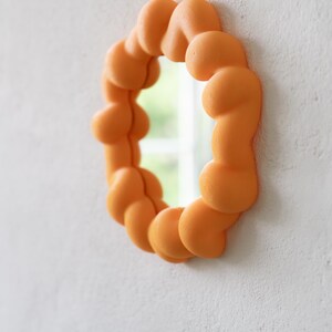 May include: An orange, oval-shaped mirror with a unique, bubbly frame. The frame is made of rounded, interconnected shapes, creating a textured appearance. The mirror is mounted on a white wall, reflecting a glimpse of the outdoors.