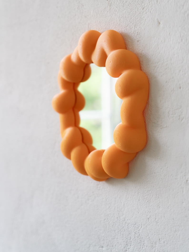May include: An orange, oval-shaped mirror with a unique, bubbly frame. The frame is made of rounded, interconnected shapes, creating a textured appearance. The mirror is mounted on a white wall, reflecting a glimpse of the outdoors.