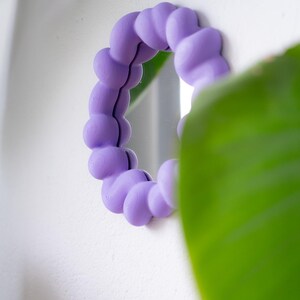May include: A round, small mirror with a lavender-colored, bubble-shaped frame. The mirror is mounted on a white wall, with a green plant partially obscuring the view. The frame's design adds a unique, playful touch to the decor.
