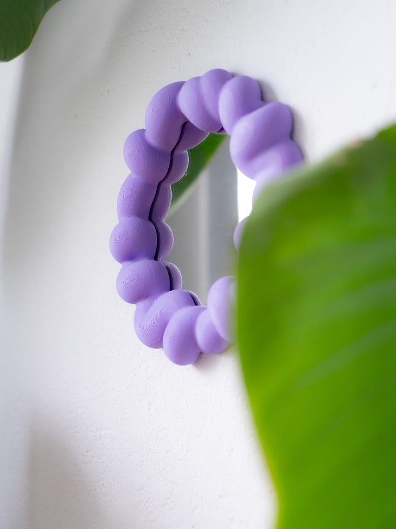 May include: A round, small mirror with a lavender-colored, bubble-shaped frame. The mirror is mounted on a white wall, with a green plant partially obscuring the view. The frame's design adds a unique, playful touch to the decor.