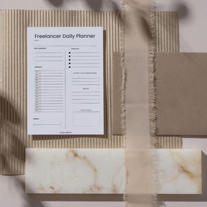 Könnte beinhalten: Ein minimalistischer Freelancer Daily Planner auf strukturiertem Hintergrund. Der Planer bietet Bereiche für Prioritäten, To-Do-Listen und Notizen. Ein marmorähnlicher Block und ein Band ergänzen die neutrale Ästhetik des Bildes.