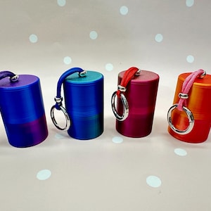 May include: Four cylindrical metal containers with matching colored cords and silver-toned key rings. The containers are arranged in a row and feature a smooth finish in shades of purple, blue, magenta, and orange, each with a secure screw-top lid.