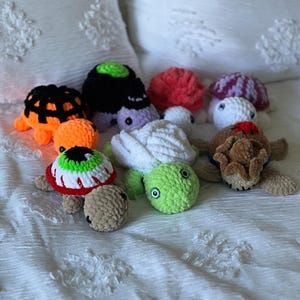May include: A collection of handmade crochet turtles in various colors and designs. The turtles have different colored shells, including orange, black, white, and green. Some turtles have unique eye designs, and the turtles are displayed on a white textured surface.