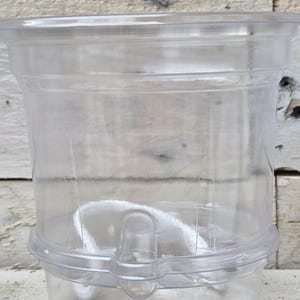 May include: A transparent plastic container with a cylindrical form. It has a broad opening at the top and a narrower base. The container includes a handle-like projection on the side, suitable for holding liquids or other items.