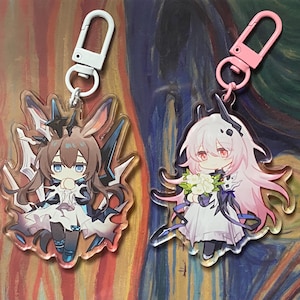 May include: Two anime-style acrylic keychains. One features a brown-haired character with blue accents and a white clip. The other has a pink-haired character with a pink clip. Both have detailed character designs and are attached to metal clasps.