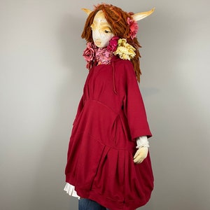 May include: A red, oversized sweatshirt with a gathered skirt-like hem. The sweatshirt is worn by a doll with a white face, red hair, and a floral crown.