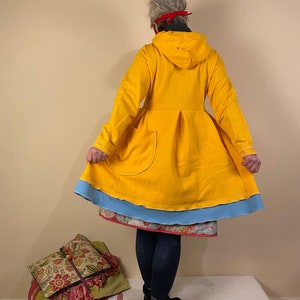 Puede incluir: Una chaqueta amarilla con capucha y ribete azul y un solo bolsillo. La chaqueta se lleva sobre una falda de estampado floral y unos vaqueros azules.