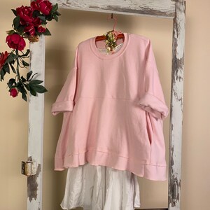 May include: An oversized pink sweatshirt with a white undershirt, hanging on a wooden door frame with a pink hanger.