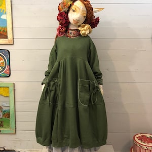 May include: A green, long-sleeved, oversized dress with pockets and a gathered skirt. The dress is worn by a doll with a white face, red hair, and a floral crown.