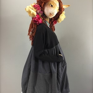 May include: A black and gray oversized tunic with a gathered skirt and pockets. The tunic is worn by a doll with red hair and a floral crown.