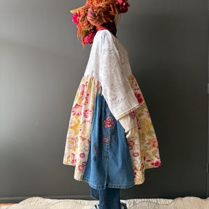 May include: A unique, handmade outfit on a mannequin. The outfit features a white floral patterned top, a denim skirt with embroidered details, and a colorful floral patterned skirt. The mannequin is adorned with a whimsical headpiece with flowers and a feather.