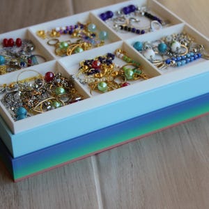 May include: A rectangular white jewelry organizer tray with six compartments filled with assorted necklaces, bracelets, and earrings featuring colorful beads and metallic chains. The tray sits inside a stacked blue and green gradient base.
