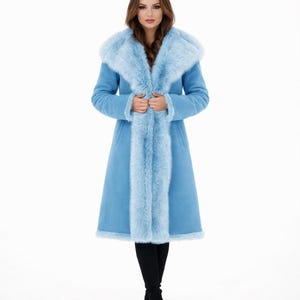 May include: A long, pale blue coat featuring a faux fur collar and trim. The coat has a tailored fit and reaches the knees. The faux fur trim is a lighter shade of blue, creating a contrast. The coat is worn over black trousers.