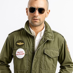 May include: A man wearing gold-framed aviator sunglasses and an olive green military-style jacket. The jacket has a white button with the text "We Are The People" and a patch with a gold wing design.