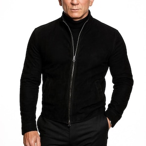 Pode incluir: Casaco preto de camurça com zíper completo, usado sobre uma gola alta preta. O casaco tem dois bolsos laterais e é combinado com calças pretas. A pessoa está em pé contra um fundo branco.