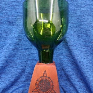 May include: A green glass goblet with a wide bowl and a narrow stem, mounted on a reddish-brown wooden base. The base features a carved Celtic knot design. The goblet is made from a recycled wine bottle.