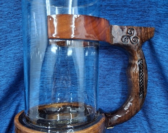 May include: A clear glass mug with a wooden base and handle. The handle is intricately carved with Celtic knot designs and a braided leather detail. The wood has a rich, dark brown finish.