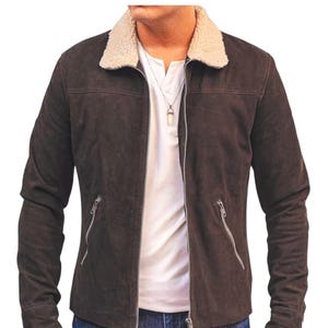 May include: A brown suede jacket with a cream-colored shearling collar. The jacket has a zip-up front, two zippered pockets, and long sleeves. The model is wearing a white shirt and blue jeans.