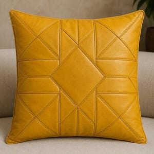 May include: A square mustard yellow throw pillow featuring a geometric pattern of stitched lines. The design consists of a central diamond shape surrounded by triangles and trapezoids, creating a symmetrical, segmented look across the entire surface.