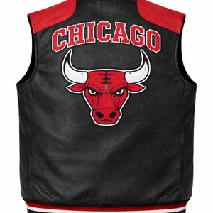 Puede incluir: Chaleco de cuero negro con detalles rojos y el logotipo de los Chicago Bulls. El chaleco presenta un dobladillo a rayas rojas y blancas y un cuello rojo. El logotipo de Chicago es rojo con contornos blancos, y el logotipo del toro es rojo con cuernos blancos.