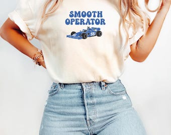 Op de afbeelding: Crèmekleurig T-shirt met de tekst "SMOOTH OPERATOR" in blauw, boven een blauwe raceauto-afbeelding. De raceauto heeft het nummer "55". Het shirt wordt gecombineerd met lichtblauwe jeans.