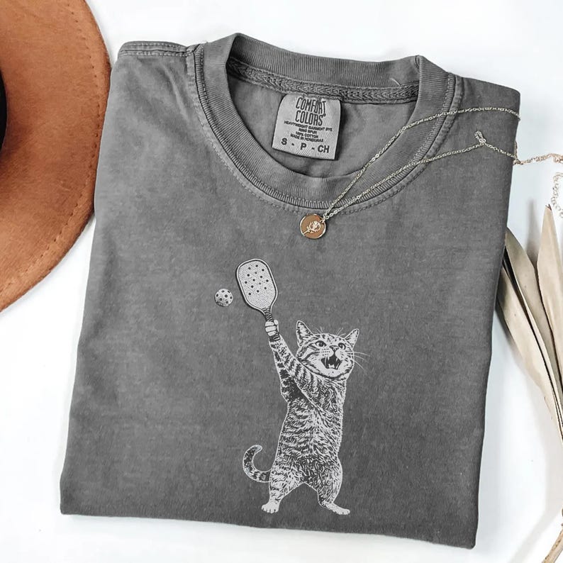 May include: A gray Comfort Colors t-shirt featuring a cat playing pickleball. The cat is holding a pickleball paddle and the ball is in the air. A silver necklace with a pendant is draped on the shirt.