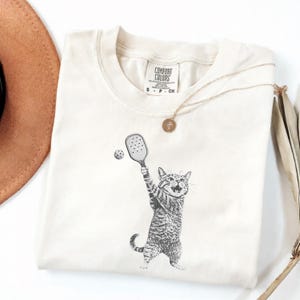 May include: Cream-colored t-shirt featuring a black and white illustration of a cat playing pickleball. The cat is holding a paddle and hitting a ball. The shirt has the words "Comfort Colors" printed on the collar.