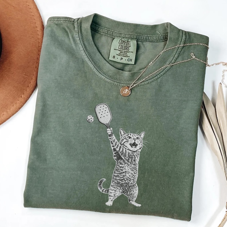 May include: Olive green Comfort Colors t-shirt featuring a graphic of a cat playing pickleball. The cat is holding a paddle and hitting a ball. A gold necklace with a pendant is also visible.
