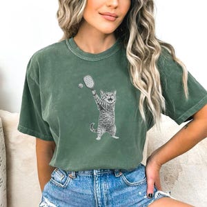 May include: Green t-shirt featuring a playful cat playing pickleball. The cat is depicted in a detailed black and white illustration, holding a paddle and hitting a ball. The shirt is a solid green color and has a relaxed fit.