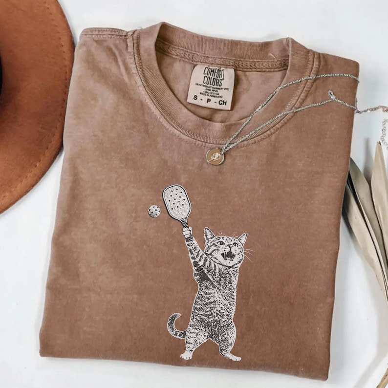 May include: Brown Comfort Colors t-shirt with a graphic of a cat playing pickleball. The cat is depicted in black and white, holding a paddle and hitting a ball. The shirt has a crew neck and the Comfort Colors logo.