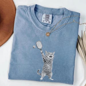 May include: A light blue t-shirt featuring a graphic of a cat playing pickleball. The cat is depicted in black and white, holding a paddle and hitting a ball. The shirt has a crew neck and the brand "Comfort Colors" is visible.
