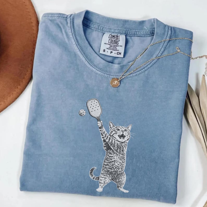 May include: A light blue t-shirt featuring a graphic of a cat playing pickleball. The cat is depicted in black and white, holding a paddle and hitting a ball. The shirt has a crew neck and the brand "Comfort Colors" is visible.