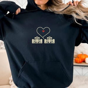 May include: Black hooded sweatshirt featuring a heart-shaped design with two race cars and a red heart. The cars are beige and connected by a white heart outline. The sweatshirt has a front pocket and a drawstring hood.