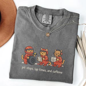 May include: A gray Comfort Colors t-shirt featuring a cartoon of three cats dressed as a pit crew. The cats are in red racing suits, with the text "pit stops, lap times, and caffeine" below the image. A silver necklace is draped across the shirt.