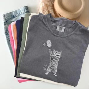 May include: A gray Comfort Colors sweatshirt featuring a cat playing pickleball. The cat is depicted with a paddle and ball. The sweatshirt is folded with other clothing items, including jeans and various colored shirts.