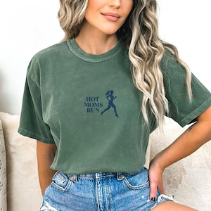 Op de afbeelding: Groen t-shirt met de tekst "HOT MOMS RUN" en een silhouet van een rennende vrouw in donkerblauw. Het shirt heeft een relaxte pasvorm en is effen groen. De tekst en de afbeelding zijn op de voorkant gedrukt.