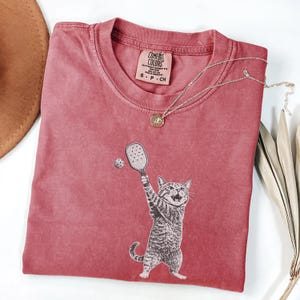 May include: A dusty rose Comfort Colors t-shirt featuring a graphic of a cat playing pickleball. The cat is holding a paddle and hitting a ball. A gold necklace with a pendant is draped across the shirt. The shirt has a small tag.