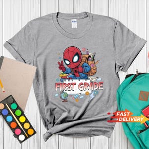 May include: A heather gray t-shirt featuring a graphic of a cartoon superhero character wearing a backpack, making a peace sign, and surrounded by school supplies like a globe, pencils, and paper with the text FIRST GRADE printed in red.