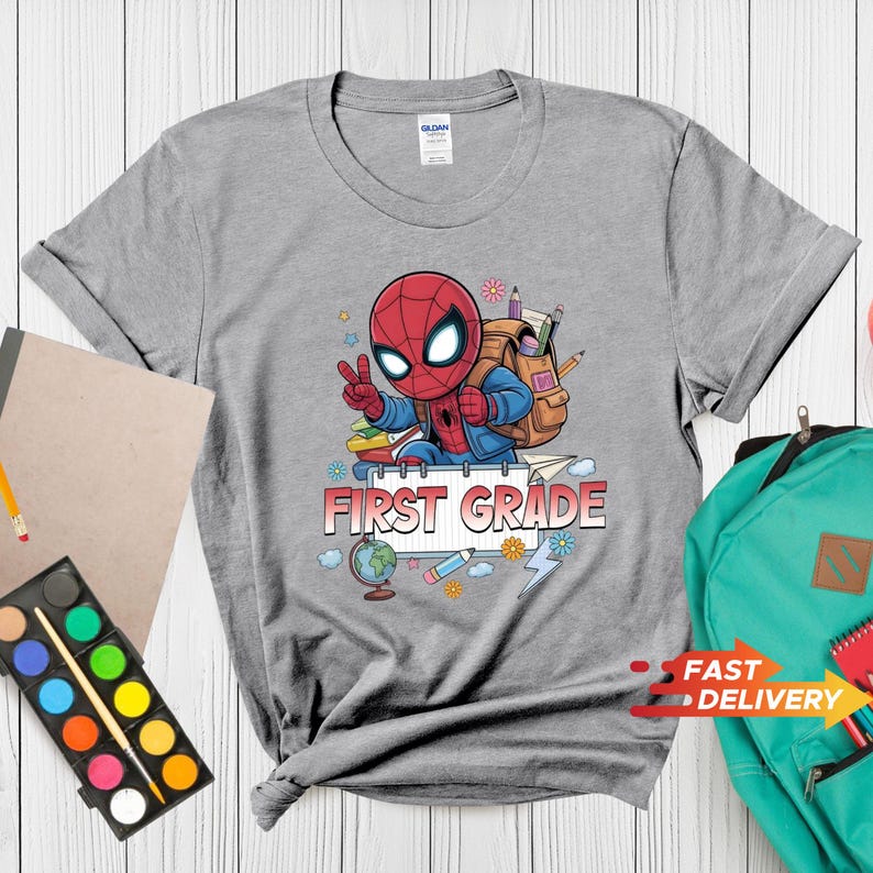 May include: A heather gray t-shirt featuring a graphic of a cartoon superhero character wearing a backpack, making a peace sign, and surrounded by school supplies like a globe, pencils, and paper with the text FIRST GRADE printed in red.